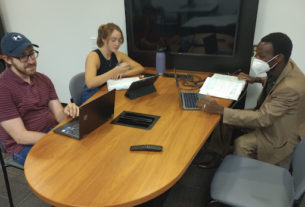 Three students sit at a table with laptops and notebooks