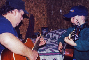 A young boy plays guitar while facing his father also playing guitar
