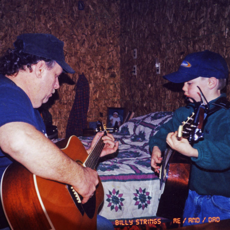 A young boy plays guitar while facing his father also playing guitar