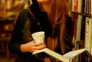 Woman smiles standing in front of a bookcase holding a book and a cup of coffee.