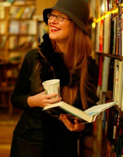 Woman smiles standing in front of a bookcase holding a book and a cup of coffee.