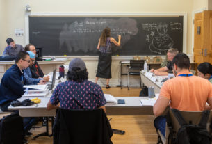 A teacher writes on the chalkboard with students sitting at tables in a U around her.