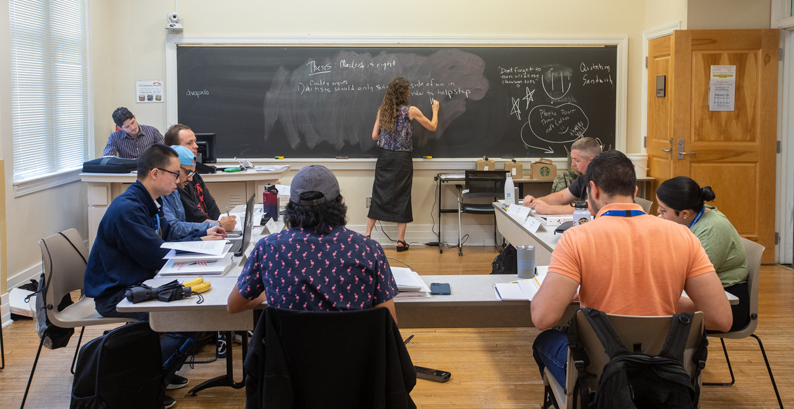 A teacher writes on the chalkboard with students sitting at tables in a U around her.