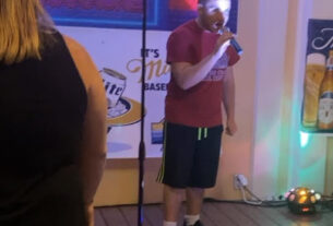 A man wearing a red shirt and black shorts sings into a microphone