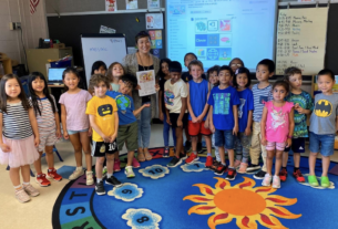 A teacher stands with her young students in her classroom, smiling at the camera