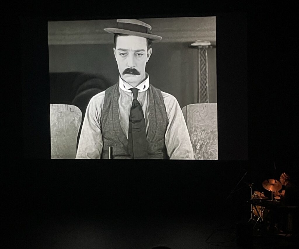 A screen Buster Keaton in "Sherlock Jr." with a man playing instruments to the bottom right of it.