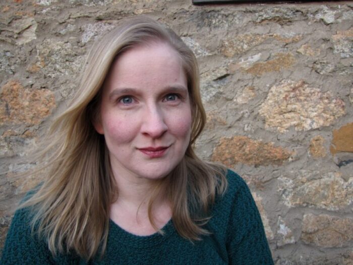 A blond woman wearing a teal sweater against a rock wall background