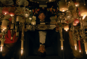 A man in a tuxedo sits at a table surrounded by candles. His reflection shows in the table.