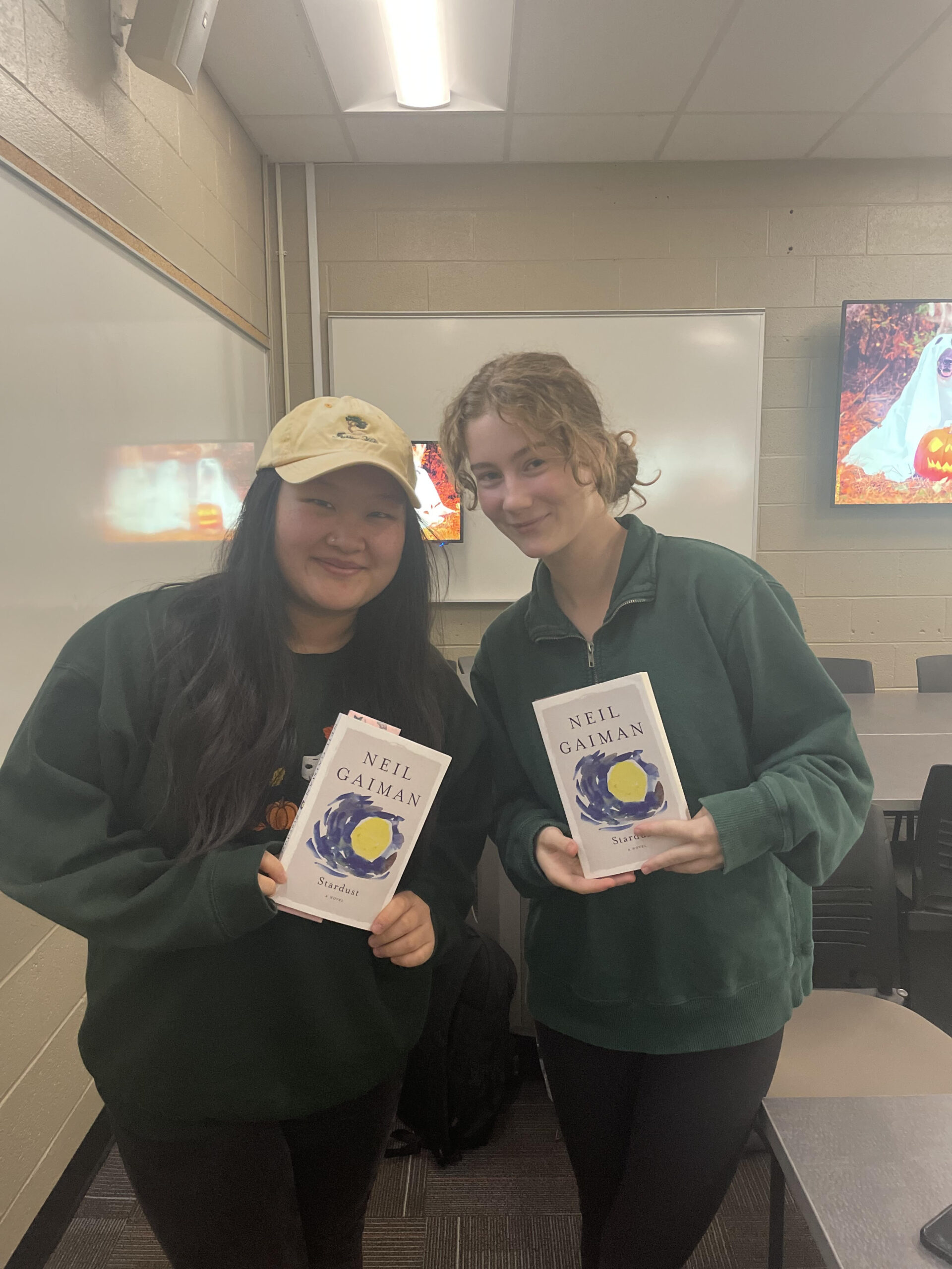Two students in green sweatshirts pose with copies of Stardust, by Neil Gaiman.