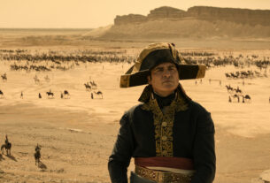 Joaquin Phoenix as Napoleon Bonaparte stands in a desert with an army on horseback in the background.