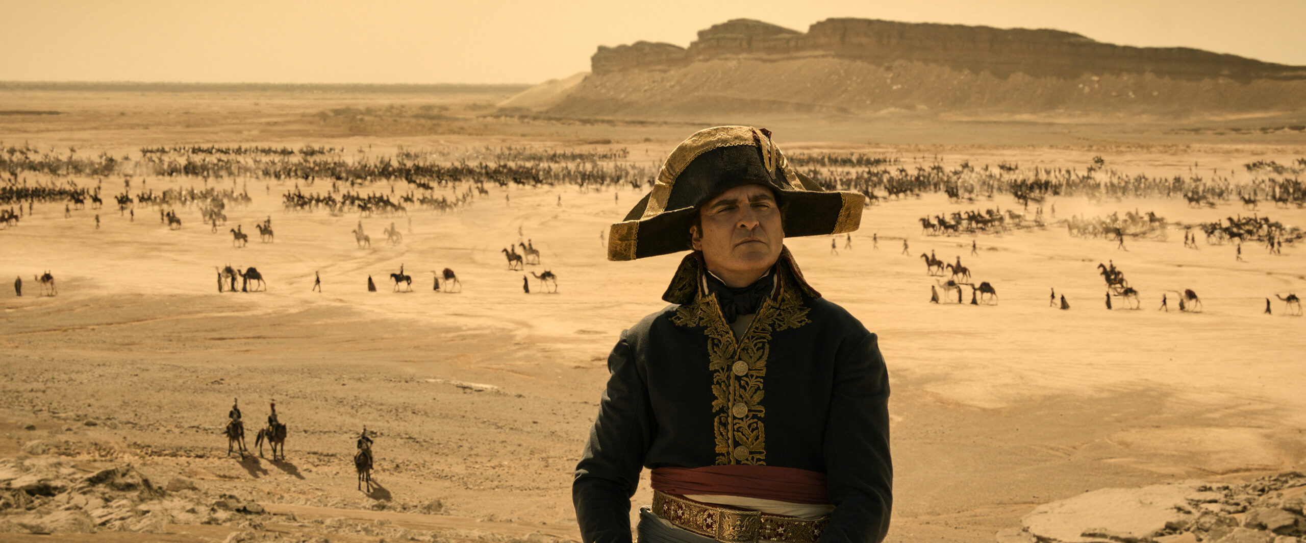 Joaquin Phoenix as Napoleon Bonaparte stands in a desert with an army on horseback in the background.