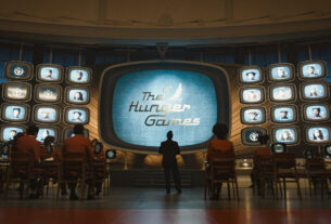 A wall of old-fashioned tv screens show the title The Hunger Games. People stand watching it.