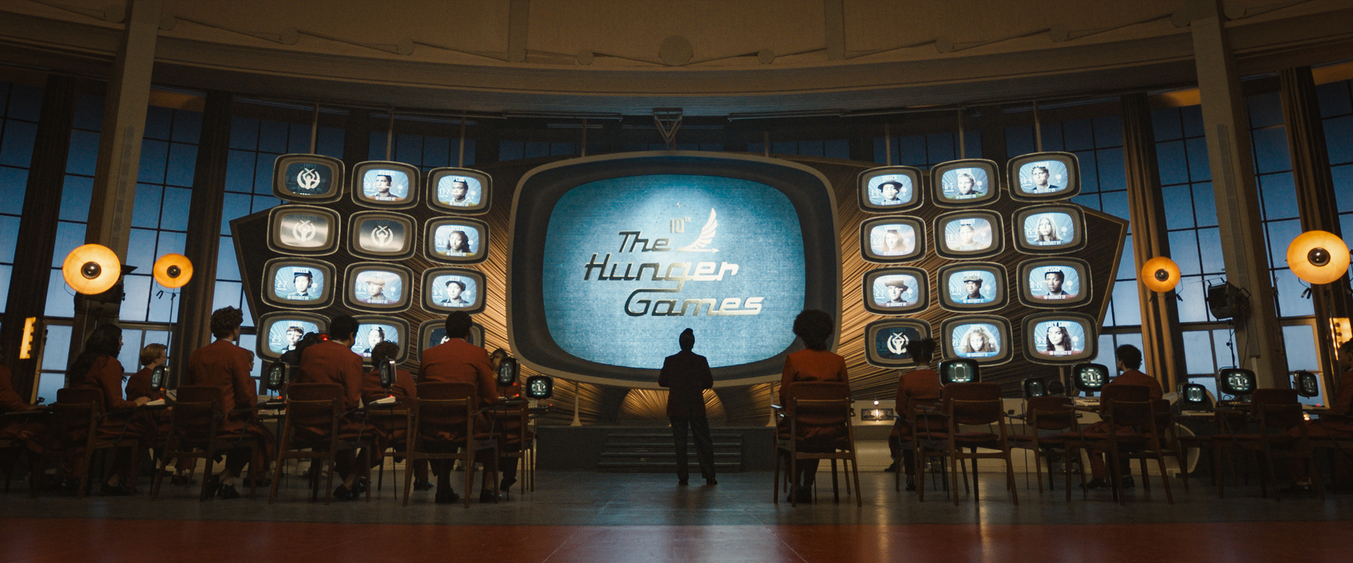 A wall of old-fashioned tv screens show the title The Hunger Games. People stand watching it.