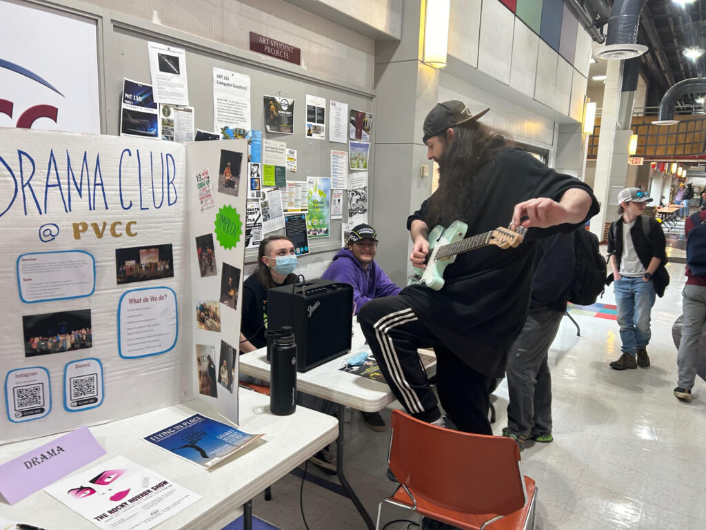 A student plays his guitar with one foot perched on a chair to another student sitting behind the display table.