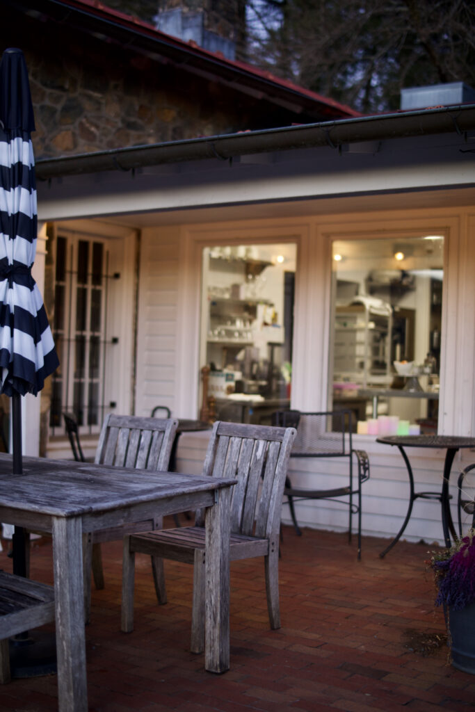 A coffee shop outdoor seating arrangement with a set of wooden chairs and table, as well as a folded striped umbrella perched in the center of the table.