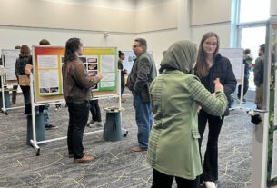 Students and guests stand talking in front of posters on whiteboards in a larger room