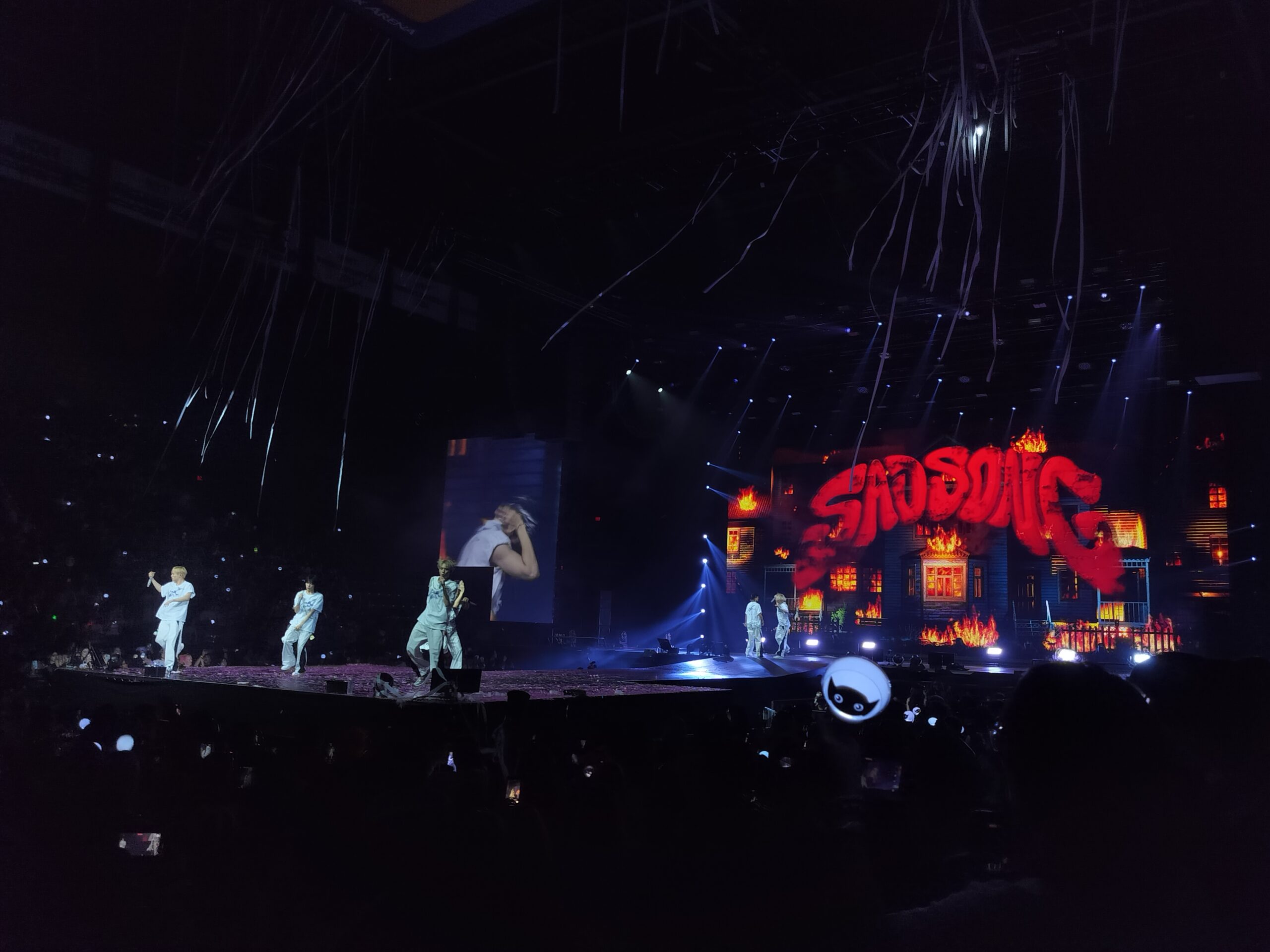 Men in white outfits sing on a stage with the words SADSONG behind them.