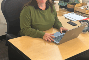 A woman in a green long-sleeved shirt smiles while at her desk with a laptop