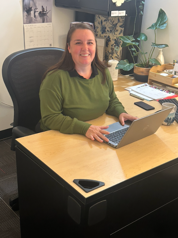 A woman in a green long-sleeved shirt smiles while at her desk with a laptop