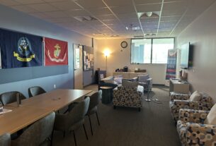 A room with a window at one end, chairs and tables, a tv screen, and military flags on the wall