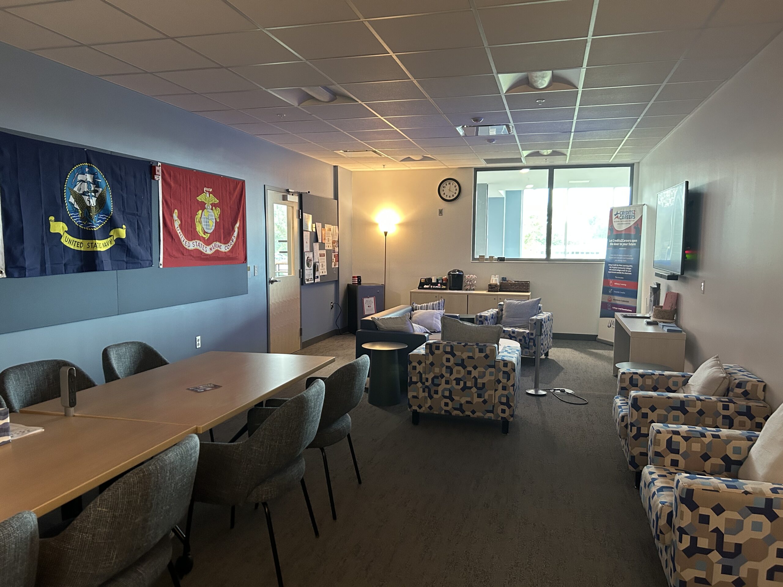 A room with a window at one end, chairs and tables, a tv screen, and military flags on the wall
