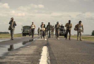 The cast of The Long Walk trudge down a damp road with military vehicles behind them