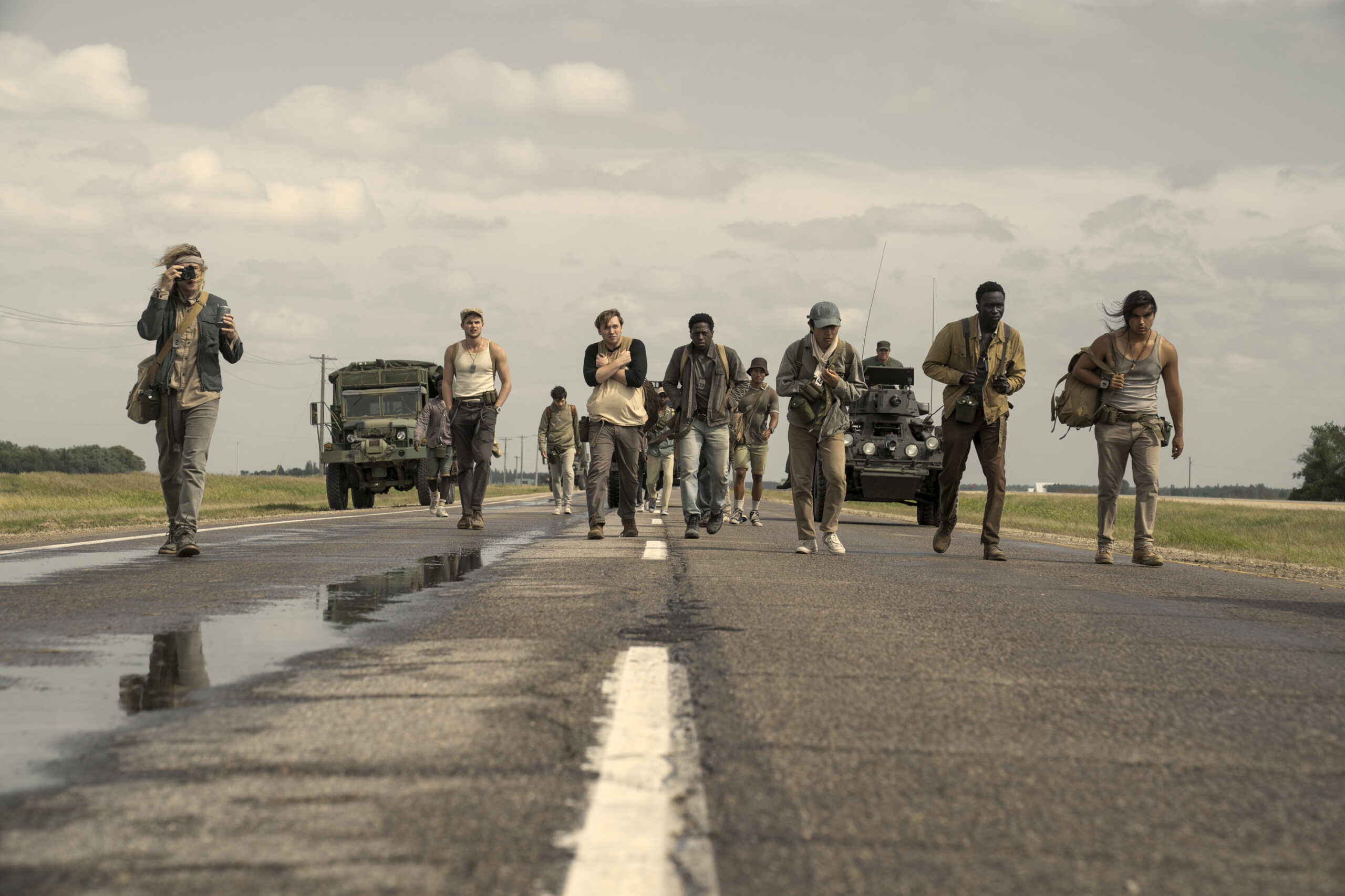 The cast of The Long Walk trudge down a damp road with military vehicles behind them