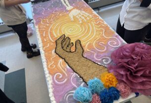 A mural made out of sugar and candy on a white table showing a skeleton hand reaching for another arm with flowers on the side and a sunset behind it.