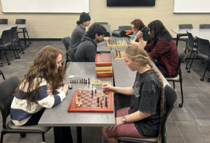 Three pairs of students play chess together at gray tables in a classroom