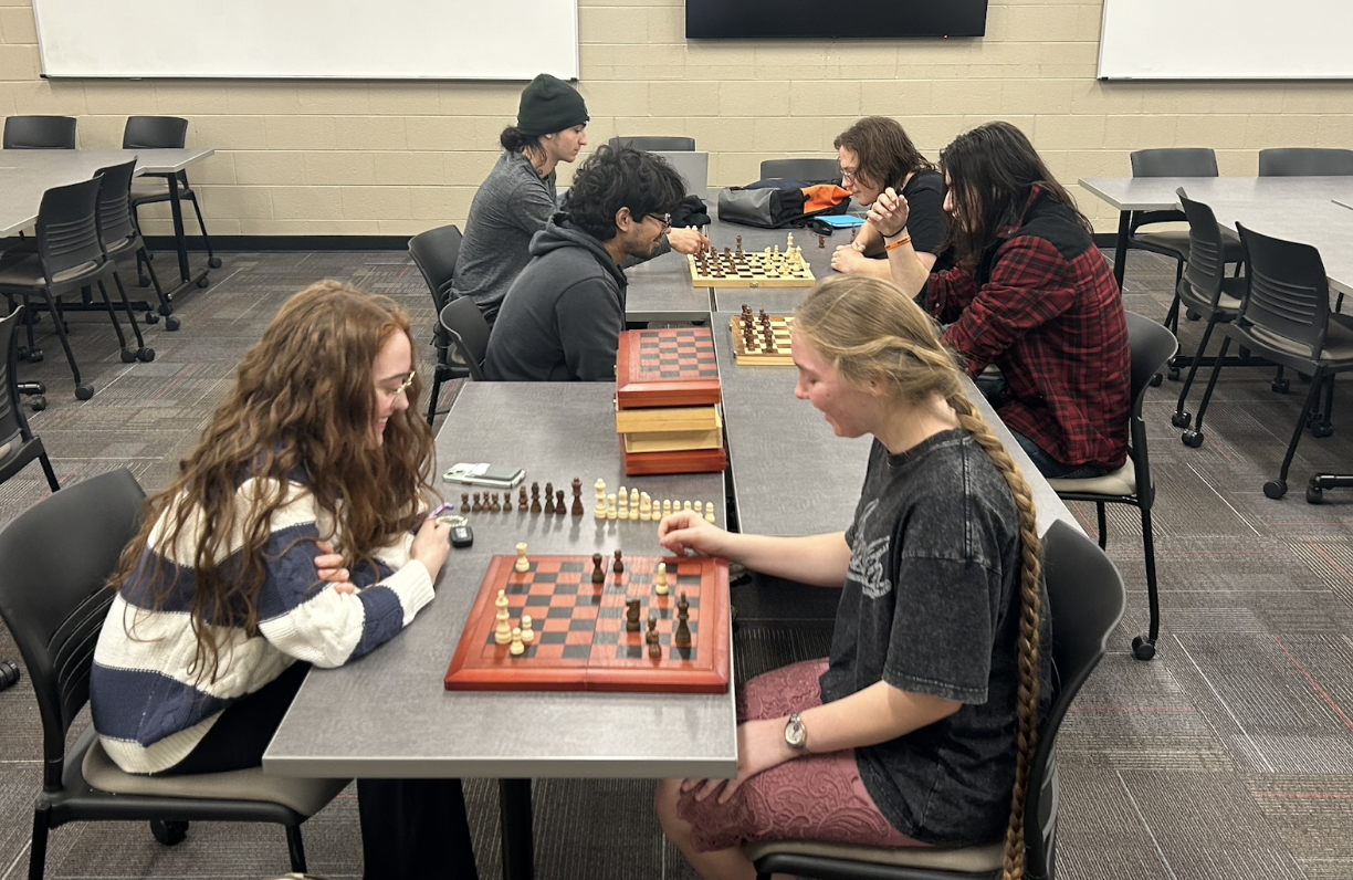 Three pairs of students play chess together at gray tables in a classroom