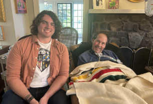 A man with curly black hair wearing a coral shirt sits next to a man under a blanket in a living room