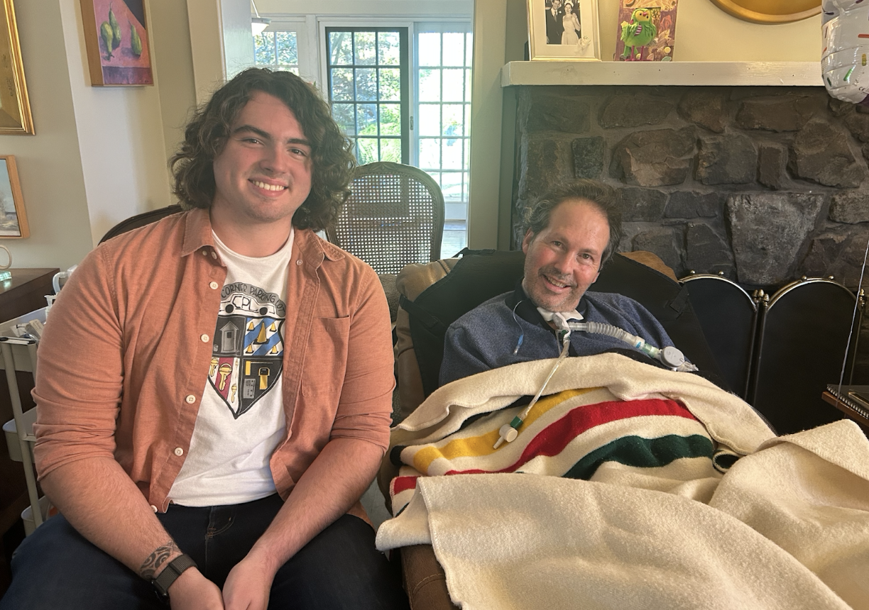 A man with curly black hair wearing a coral shirt sits next to a man under a blanket in a living room