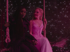 Cynthia Erivo and Ariana Grande sit together on a swing with pinpoints of light behind them against a dark background.