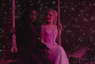 Cynthia Erivo and Ariana Grande sit together on a swing with pinpoints of light behind them against a dark background.