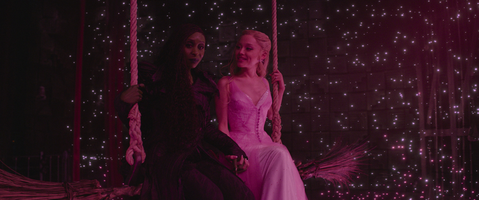 Cynthia Erivo and Ariana Grande sit together on a swing with pinpoints of light behind them against a dark background.