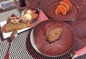 A serving of each three desserts on plates on a metal table: bread pudding with ice cream, coffee cake covered in sugar, and orange cake with two orange slices.