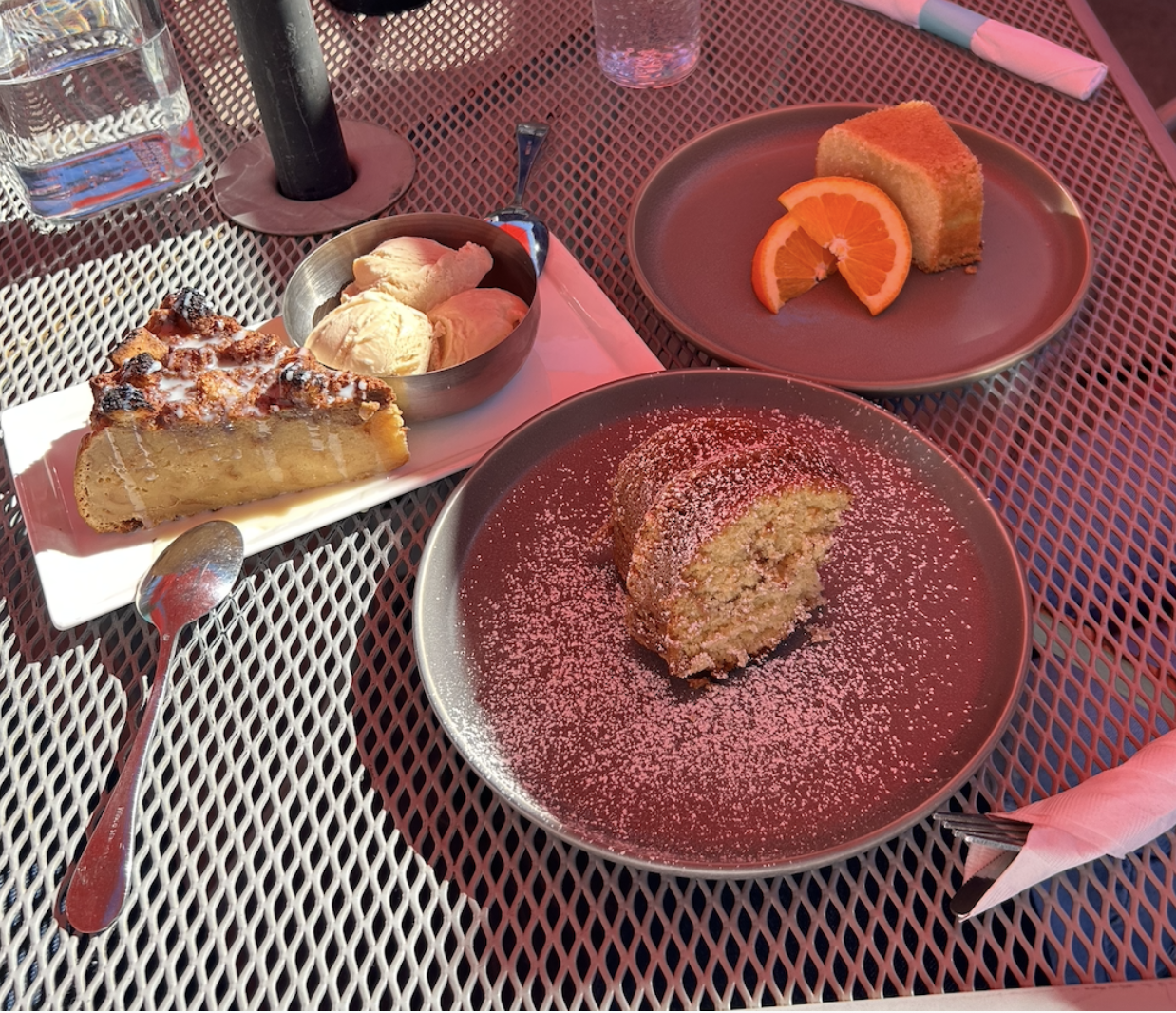 A serving of each three desserts on plates on a metal table: bread pudding with ice cream, coffee cake covered in sugar, and orange cake with two orange slices.