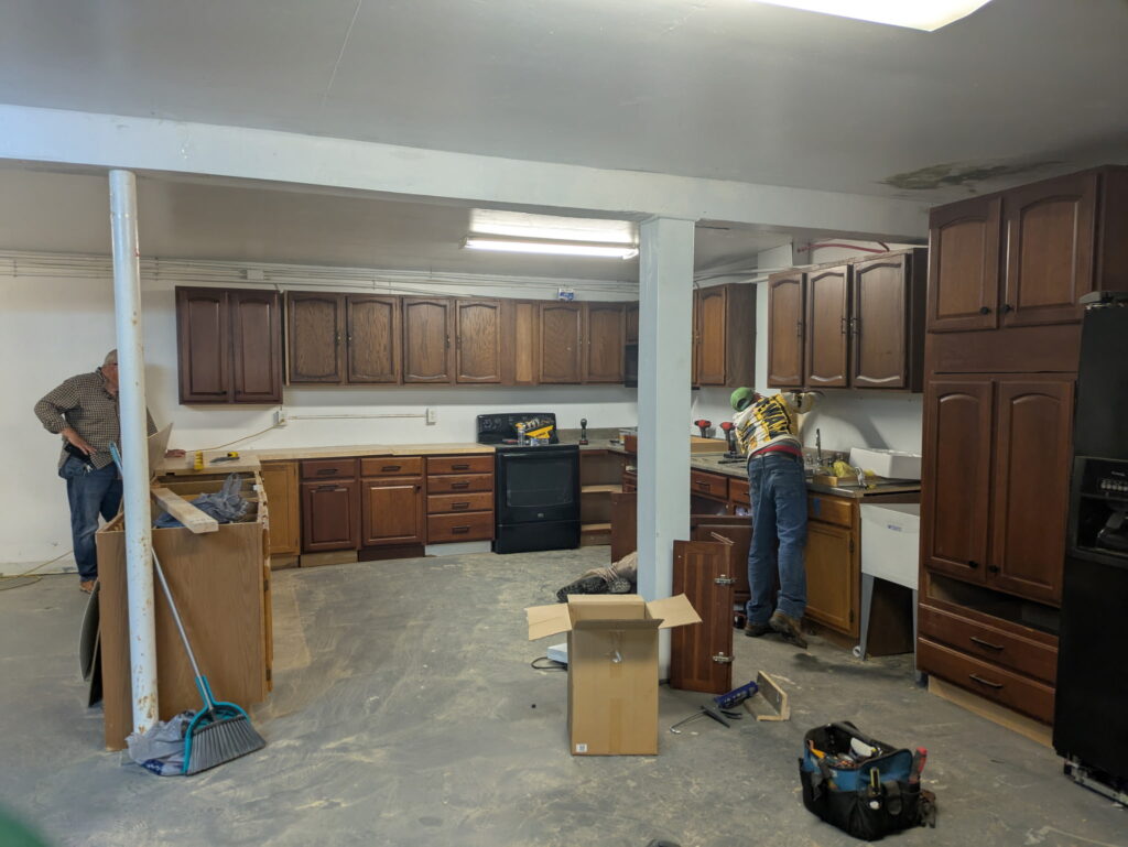 Two people work on improving a kitchen with dark wood cabinets in a church basement