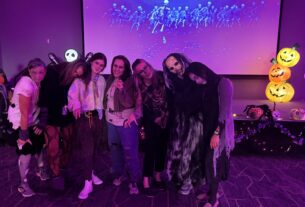 Thriller Flash Mob members pose together in zombie costumes in front of a screen showing skeletons