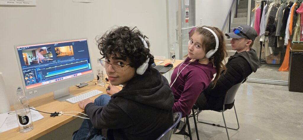 Three students sitting in front of a computer editing the movie. Two wear headphones and face the camera.
