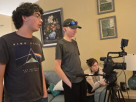 Three students behind a video camera with one holding a script.