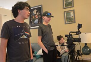 Three students behind a video camera with one holding a script.