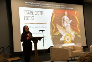Jennifer Tomás on stage with a presentation behind her, the slide titled, "History, Culture, Politics" with a picture of a historical statue of a women on the right of the slide.