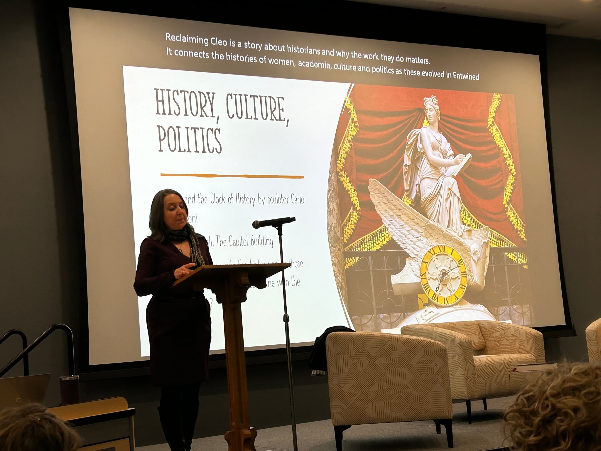 Jennifer Tomás on stage with a presentation behind her, the slide titled, "History, Culture, Politics" with a picture of a historical statue of a women on the right of the slide.