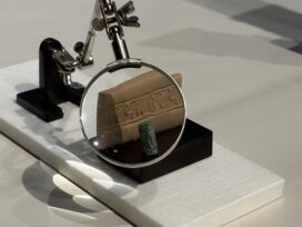 A cylindrical seal designed to be rolled across clay accompanied by a small clay tablet showcasing the seal and a magnifying glass set to magnify the tablet.