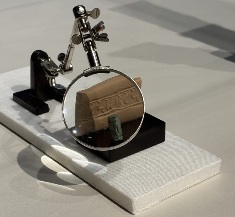 A cylindrical seal designed to be rolled across clay accompanied by a small clay tablet showcasing the seal and a magnifying glass set to magnify the tablet.