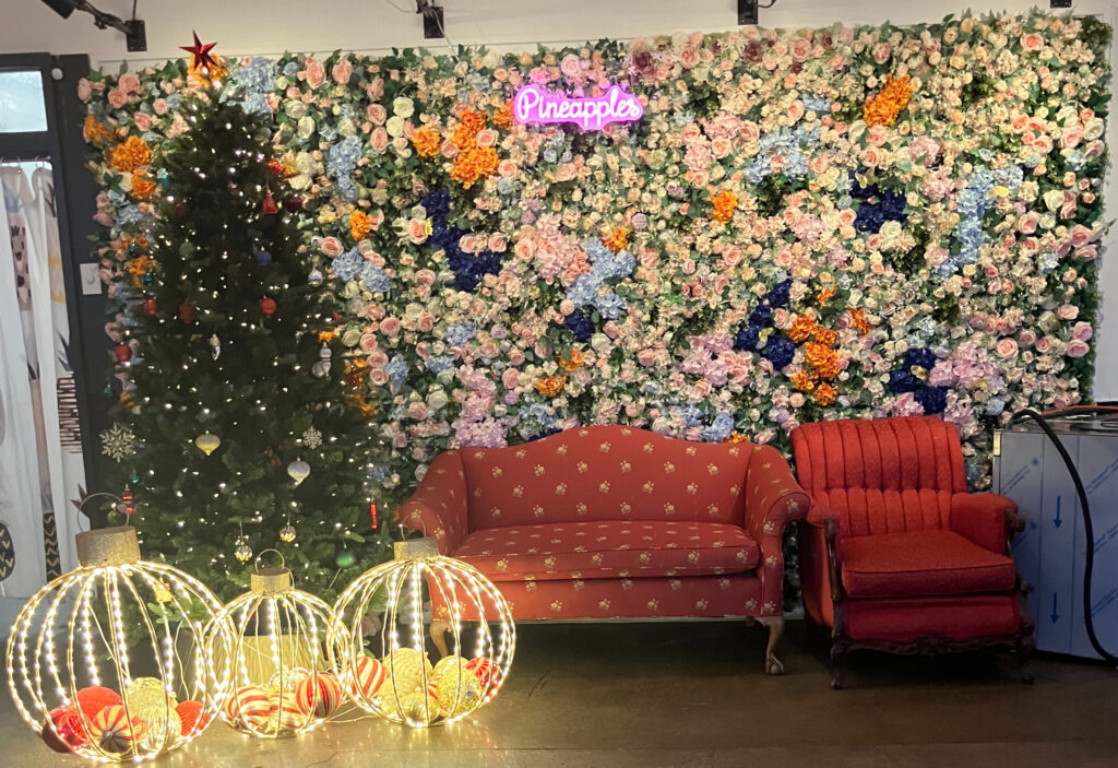 A flower wall with a neon Pineapples sign behind a red couch and chair and next to a Christmas tree and lights.