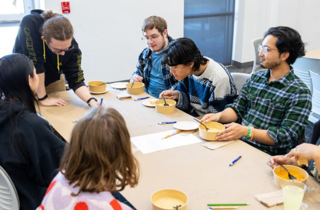 A group of students watching another student write on a table covered in paper.
