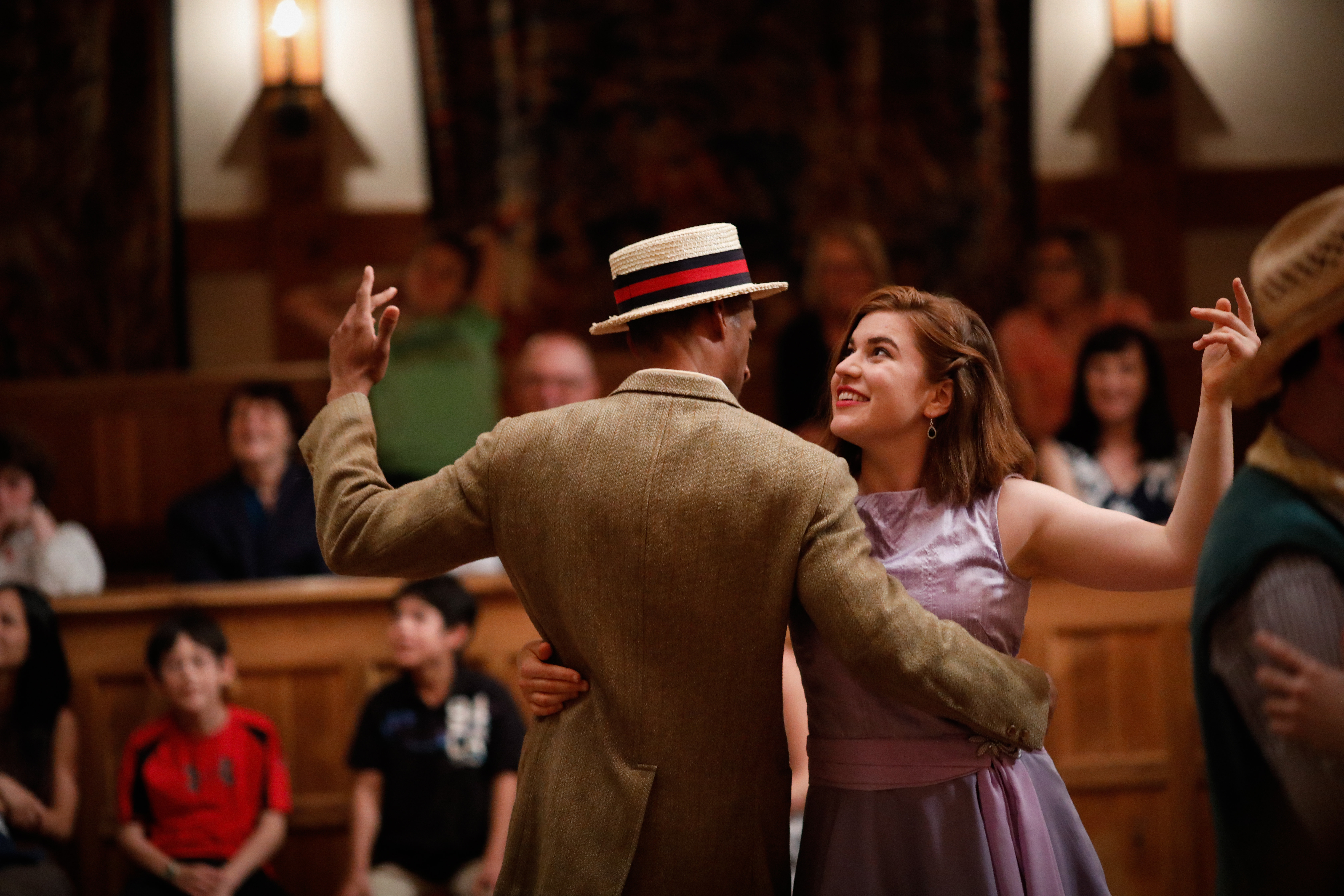 Two actors, a man in a suit and hat and a woman in a purple dress, dancing on stage in the Blackfriars Playhouse in front of an audience. 