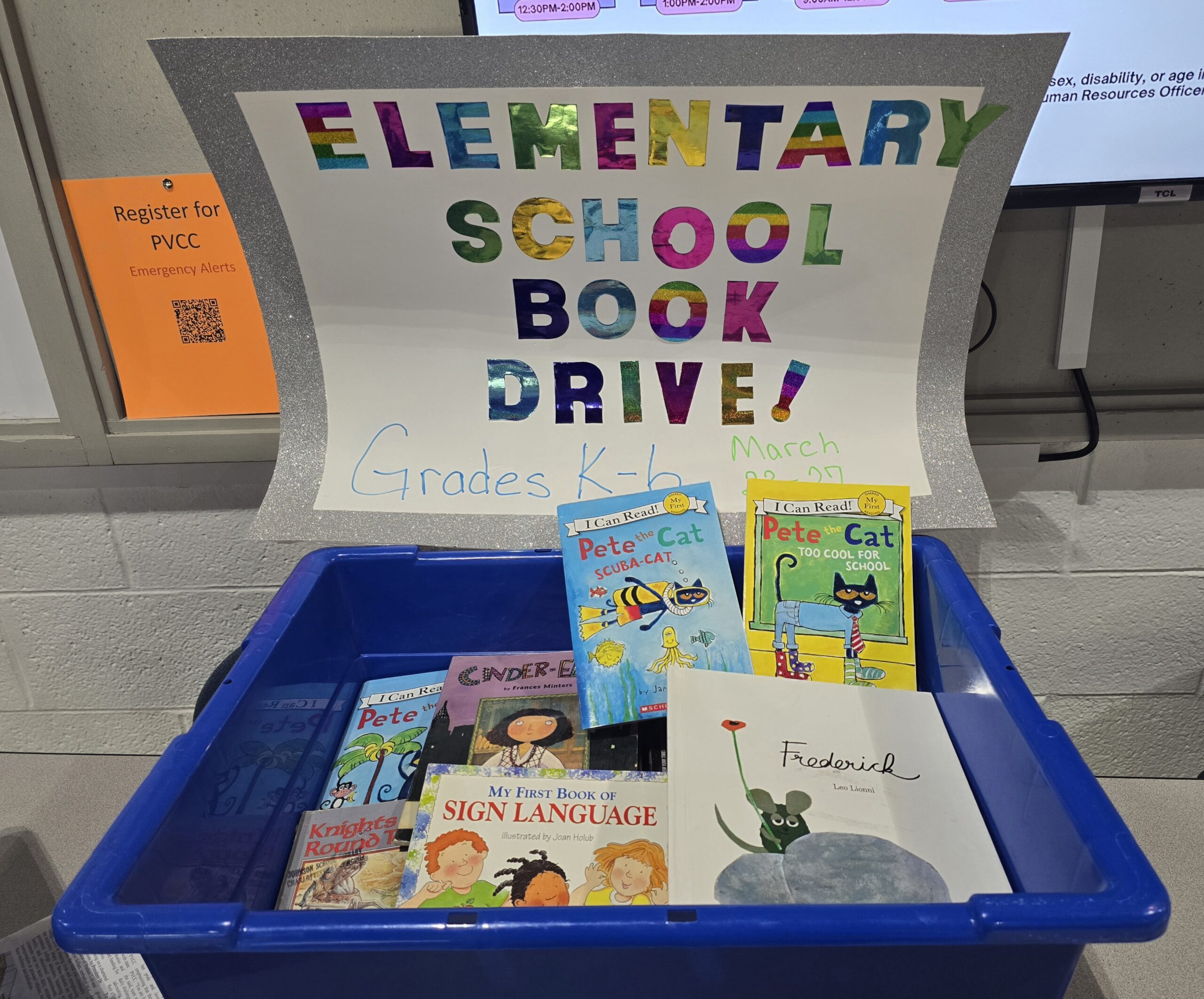 A large, blue bin with children's books in it. A big sheet of paper is connected to the top with "Elementary School Book Drive!" written on it in rainbow lettering.